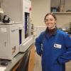 A young woman in lab gear smiles next to analytical instruments